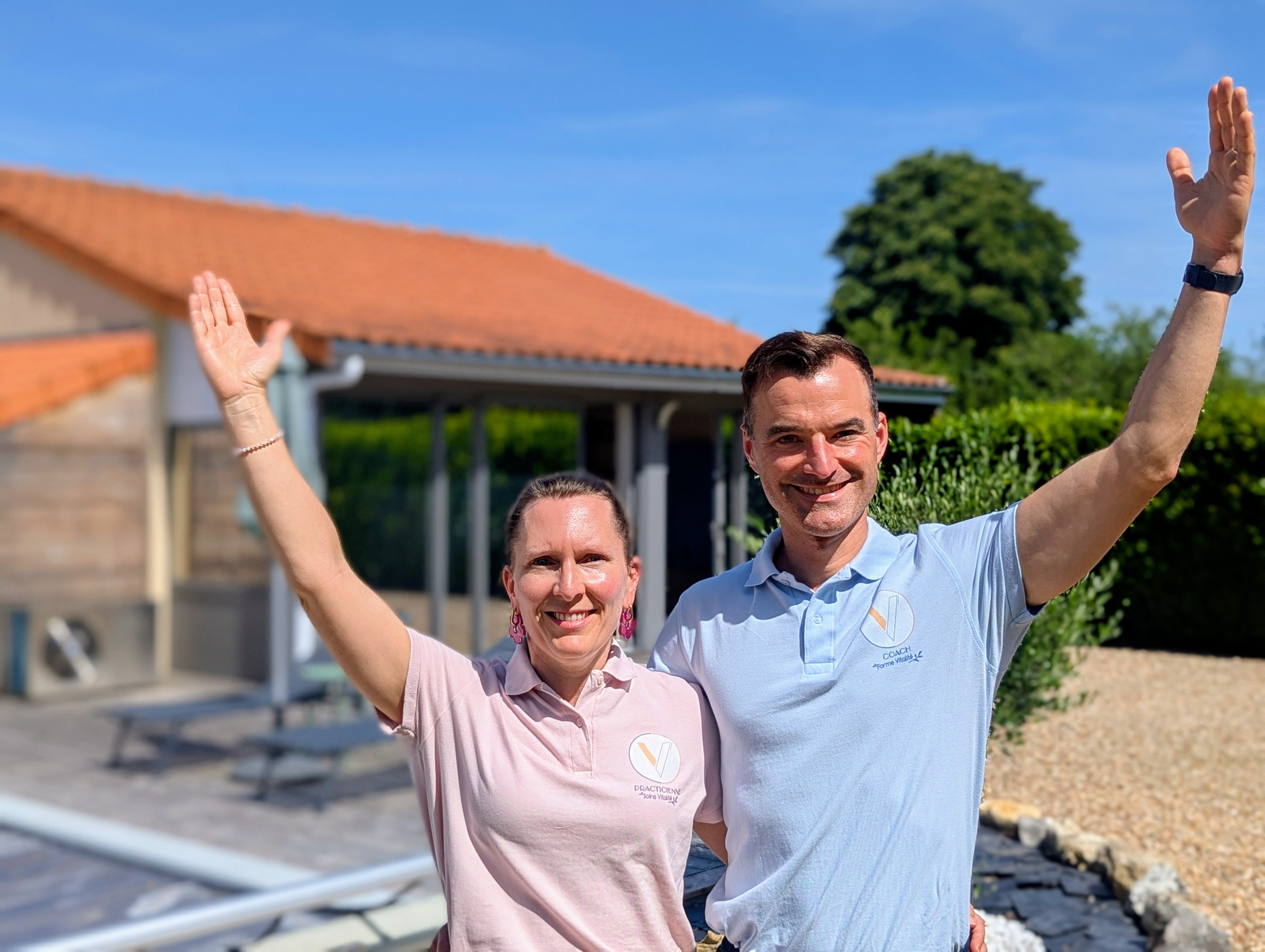Deux personnes souriantes posant devant une maison avec les mains levées en signe de salutation.