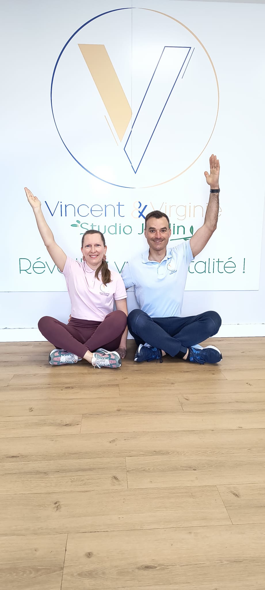 Deux personnes assises par terre, levant les bras, devant un mur avec le logo et le nom 'Vincent & Virginie - Studio Jardin'.